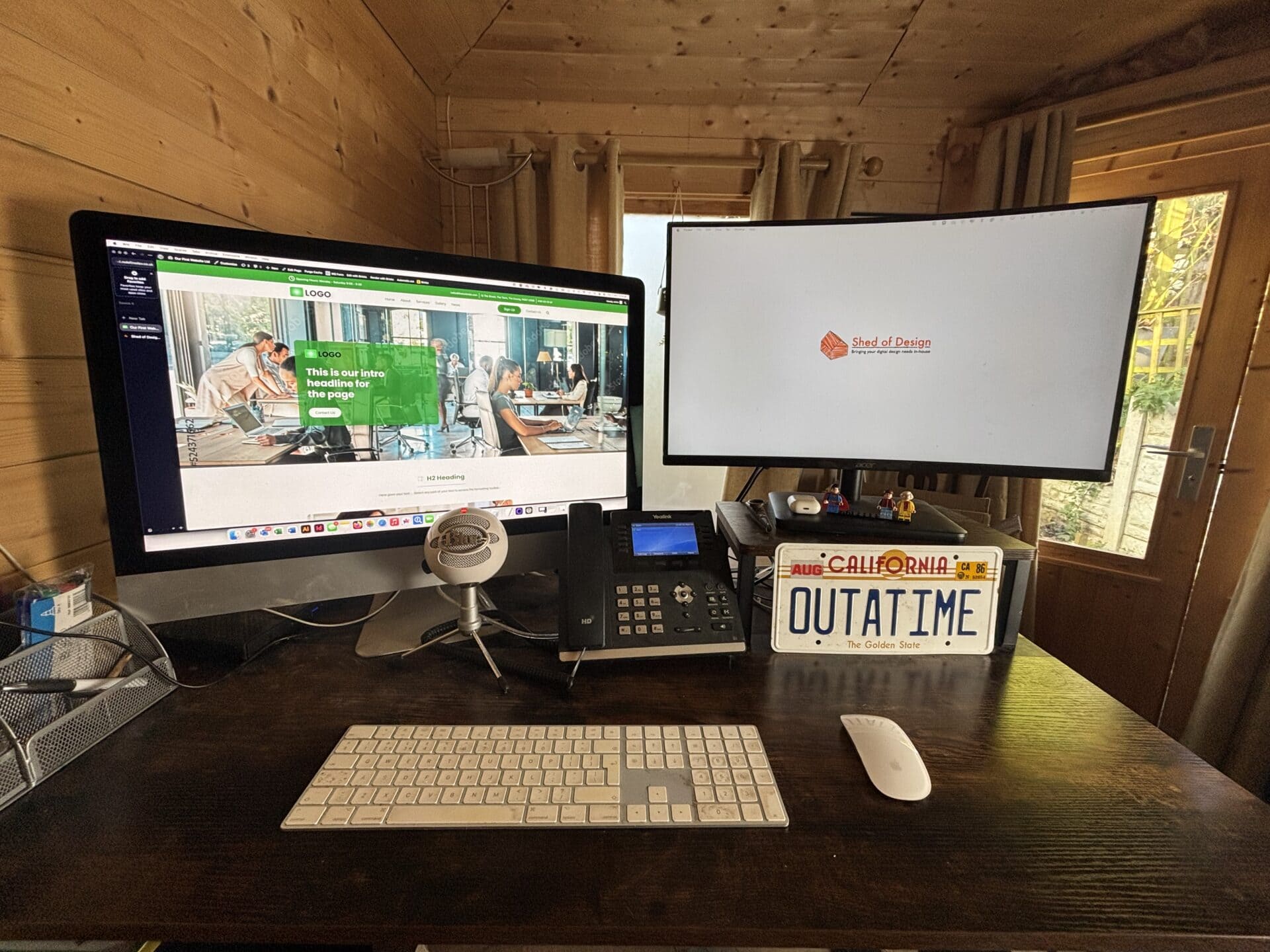A wooden home office with two monitors, a keyboard, phone, microphone, and a "OUTATIME" license plate on the desk near a window.