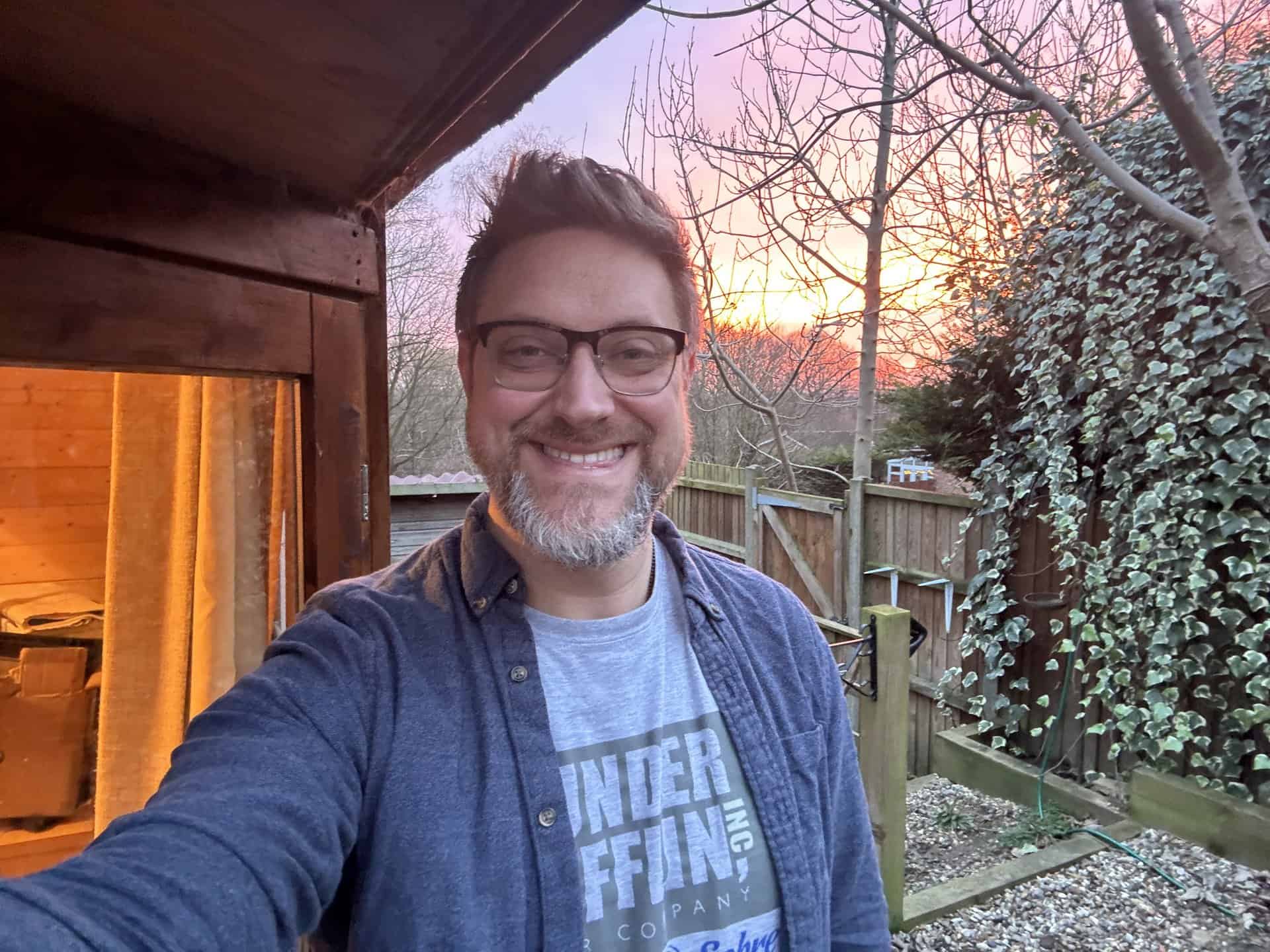 A man with glasses and a beard smiles at the camera outside near a wooden building, with trees and a sunset in the background.